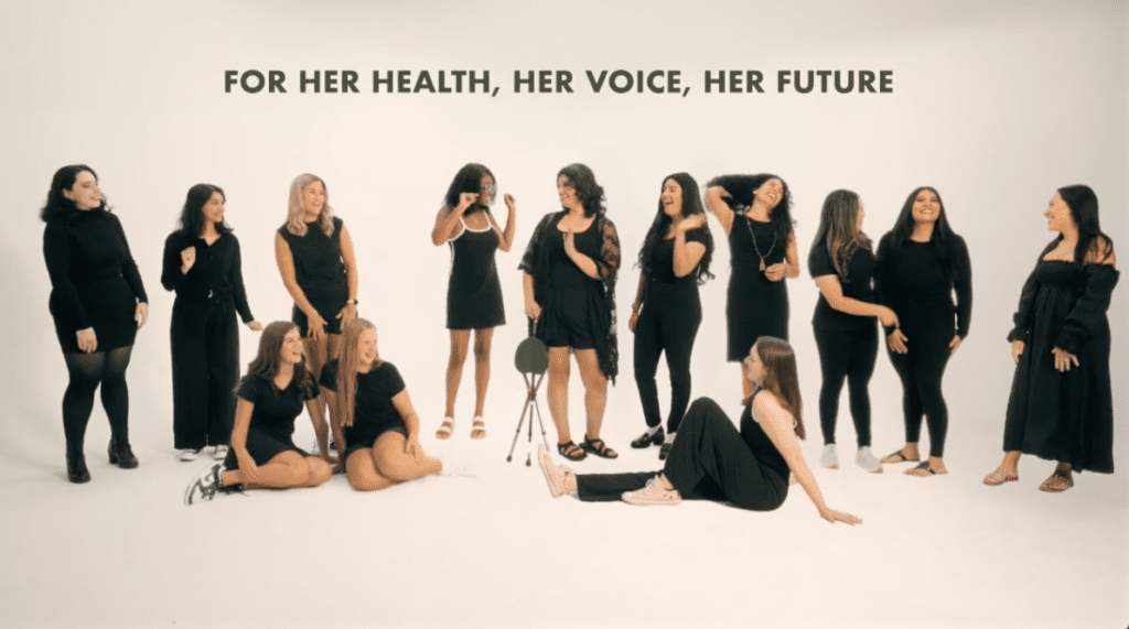 A diverse group of women stand and sit together in a bright studio setting beneath the words "For Her Health, Her Voice, Her Future," representing confidence, community, and support for women's well-being.