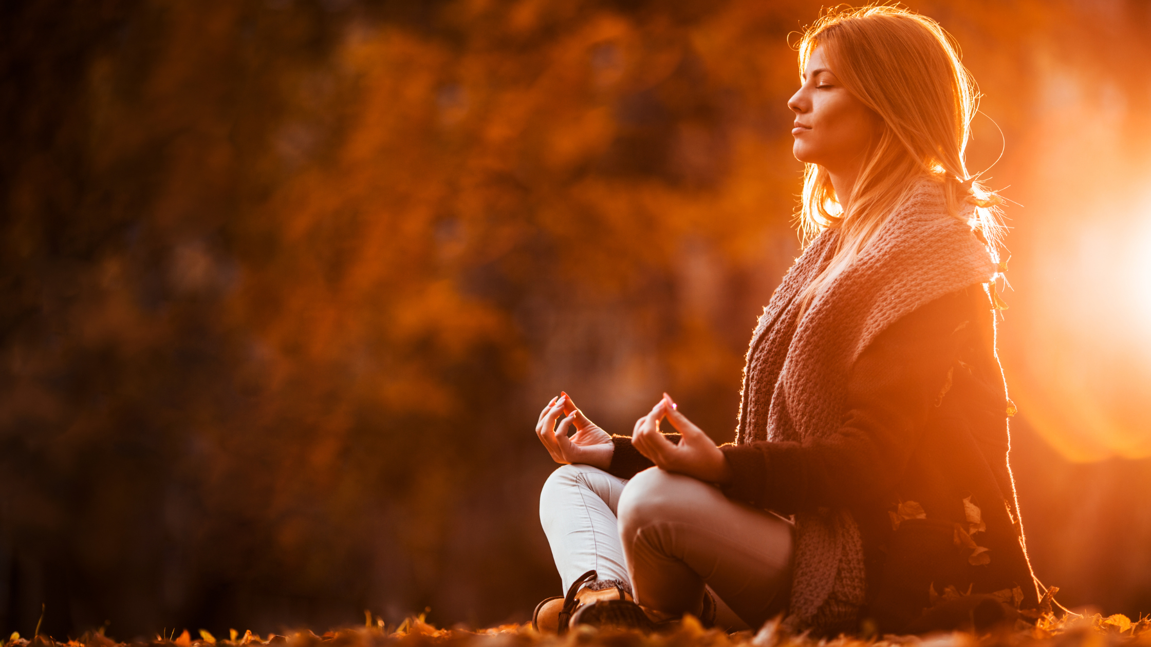 Woman sitting outdoors in autumn leaves with her eyes closed, practicing meditation in warm sunlight.