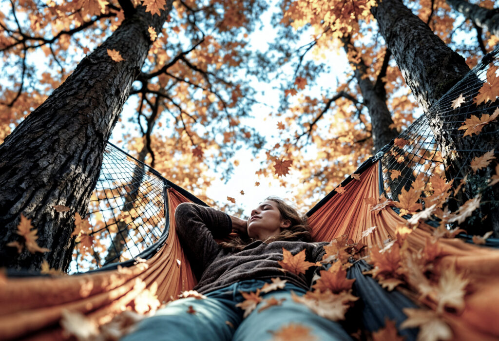 Woman relaxing in a hammock beneath autumn trees with orange leaves falling around her.