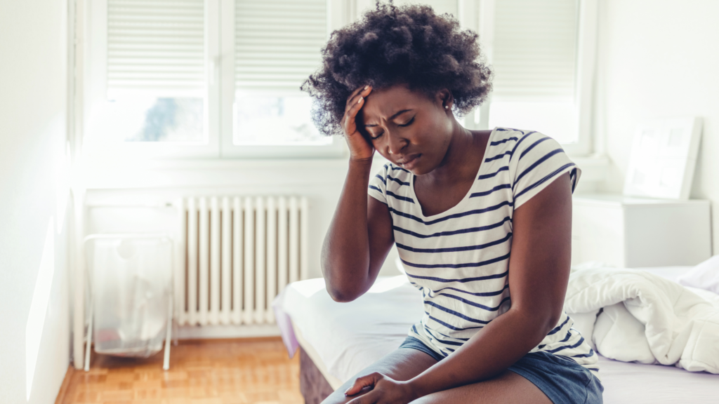 Woman sitting on the edge of a bed with her hand on her forehead, appearing stressed or overwhelmed.