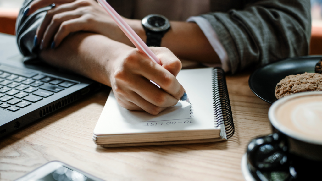 Woman writing a to-do list in a notebook beside a laptop and coffee.