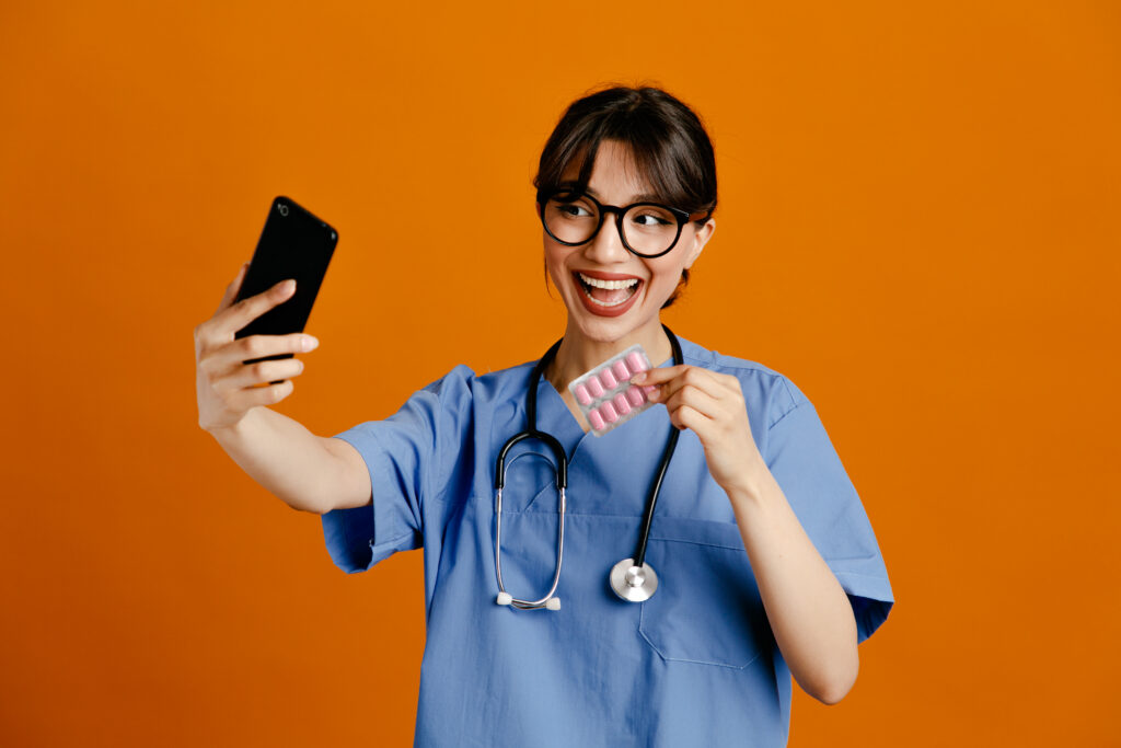 Healthcare professional in scrubs taking a selfie while holding a pack of pills.